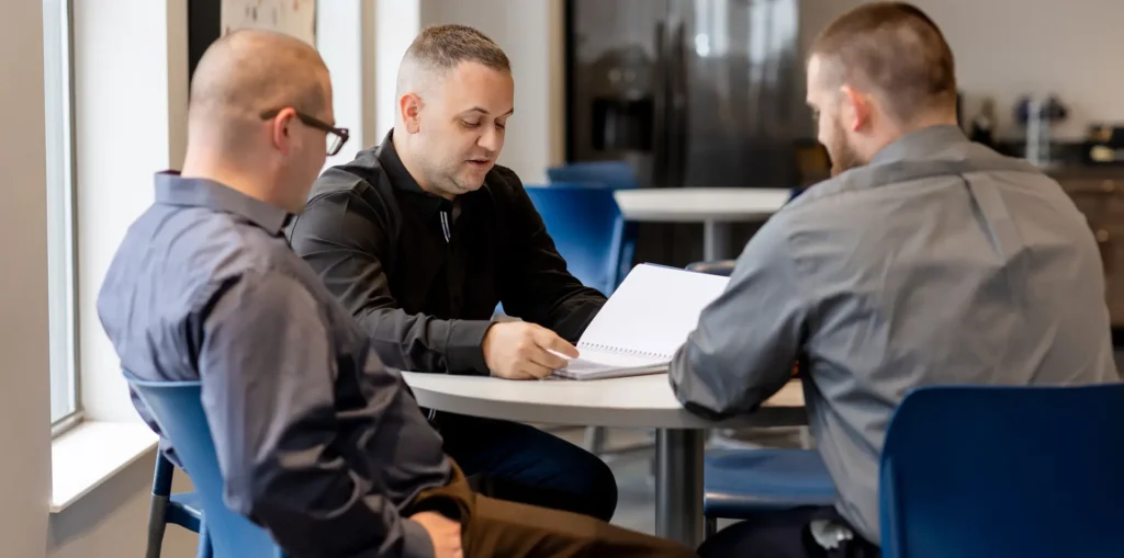 Three people working at a table discussing plans in a notebook.