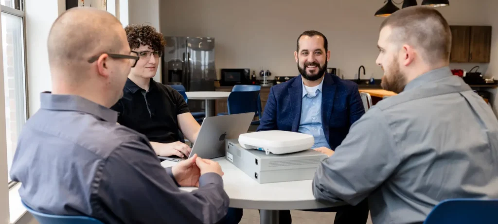 Four people sitting at a table with a laptop and network hardware.